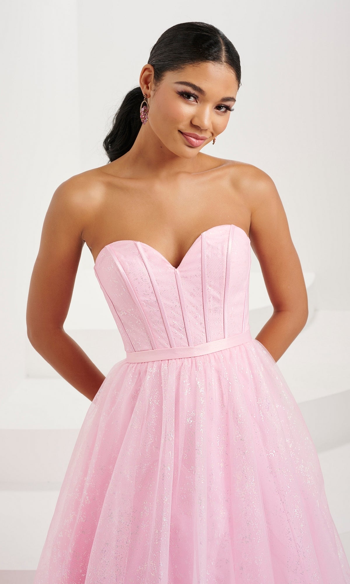 A woman poses with her hands behind her back against a plain, light-colored background, wearing the Tiffany Strapless Glitter Pastel Prom Dress 16083 featuring a shimmering corset bodice.